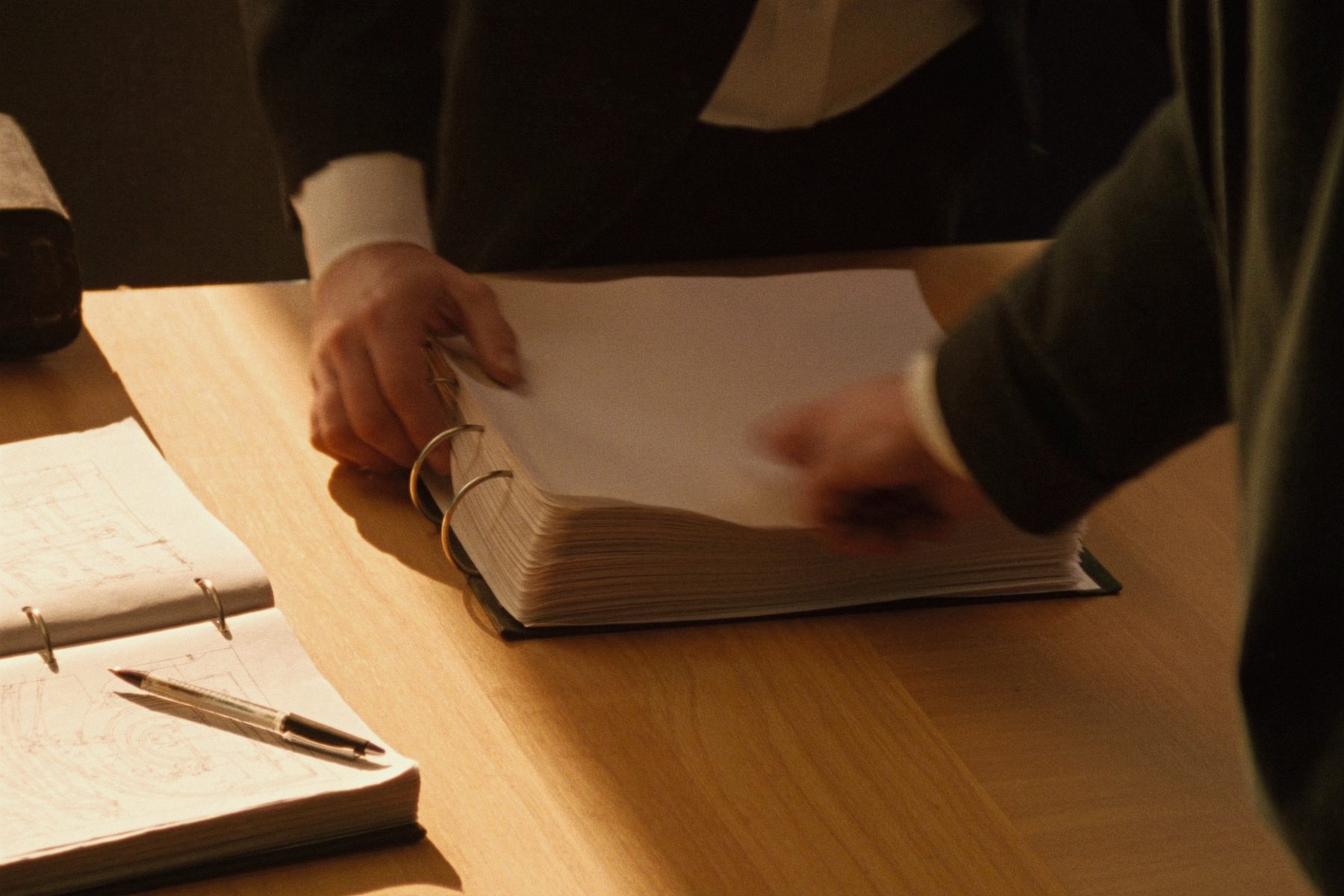 Hands turning the pages of a printed document beside a folded technical drawing in warm light.