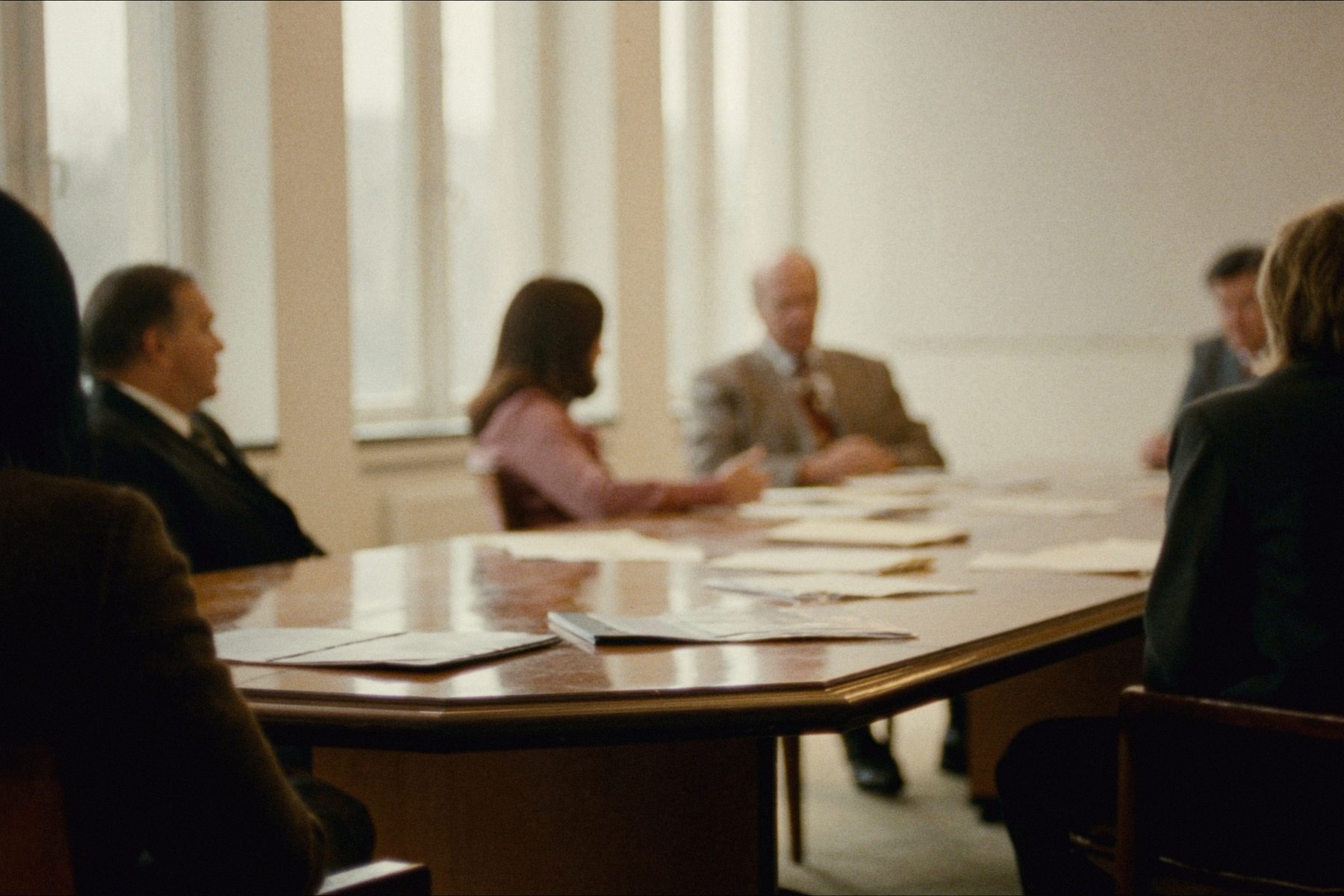A meeting across a long table in a bright room, papers and folders in front of each side.